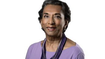 Dr. Anna Eliatamby smiling, wearing a light purple shirt and layered purple necklace, with short hair, on a plain white background.